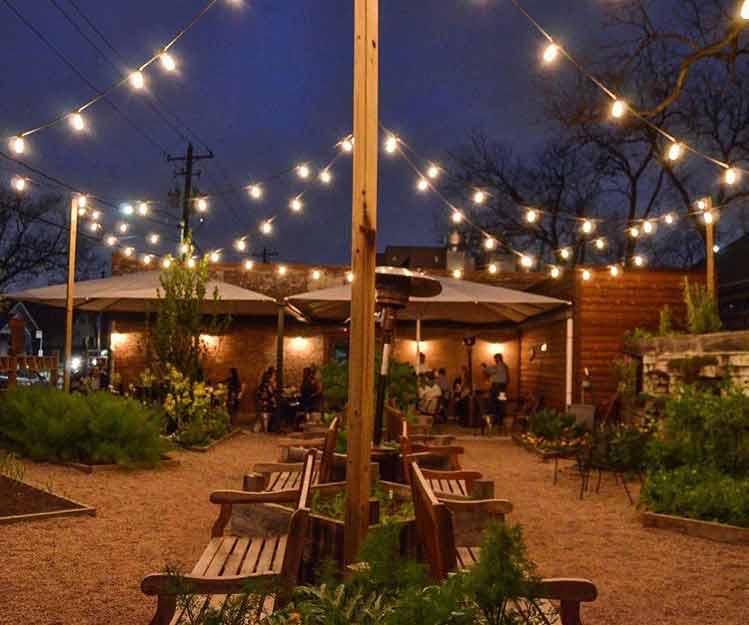 Business maintenance lighting maintains the outdoor seating area at a restauruant with wooden tables, benches, and string lights