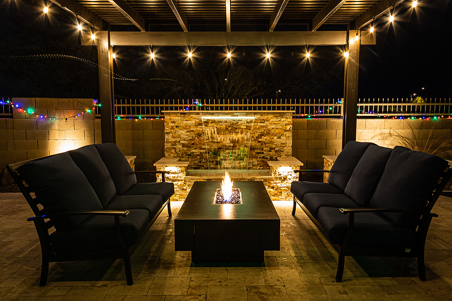 A resort style backyard at night featuring outdoor landscape lighting with a waterfall, pergola, and a firepit at night.