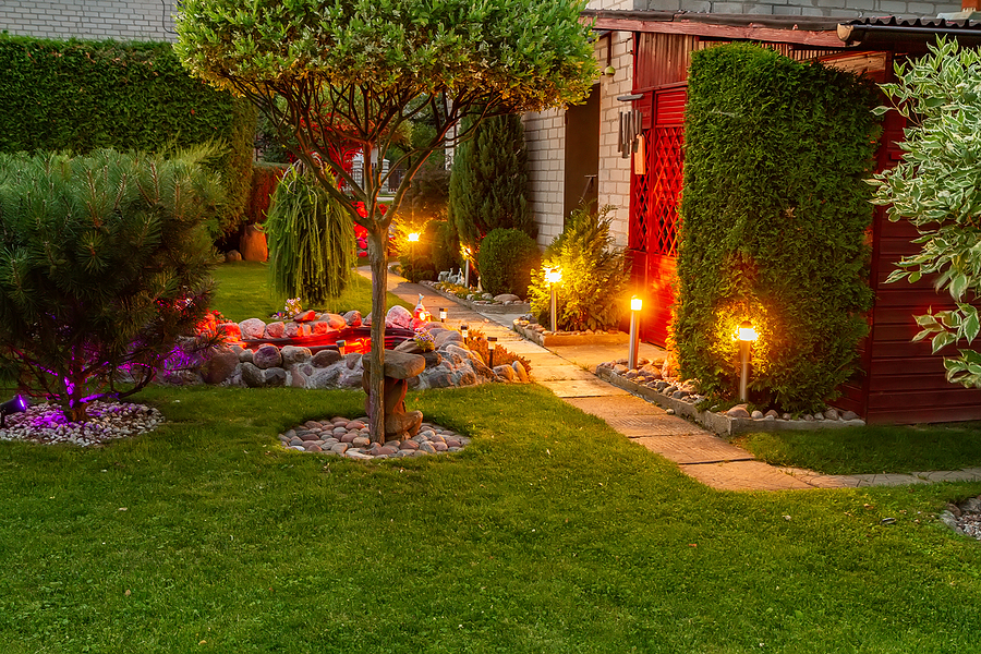 Well kept backyard with plants, trees, and bushes illuminated by path lighting leading to a magic garden.